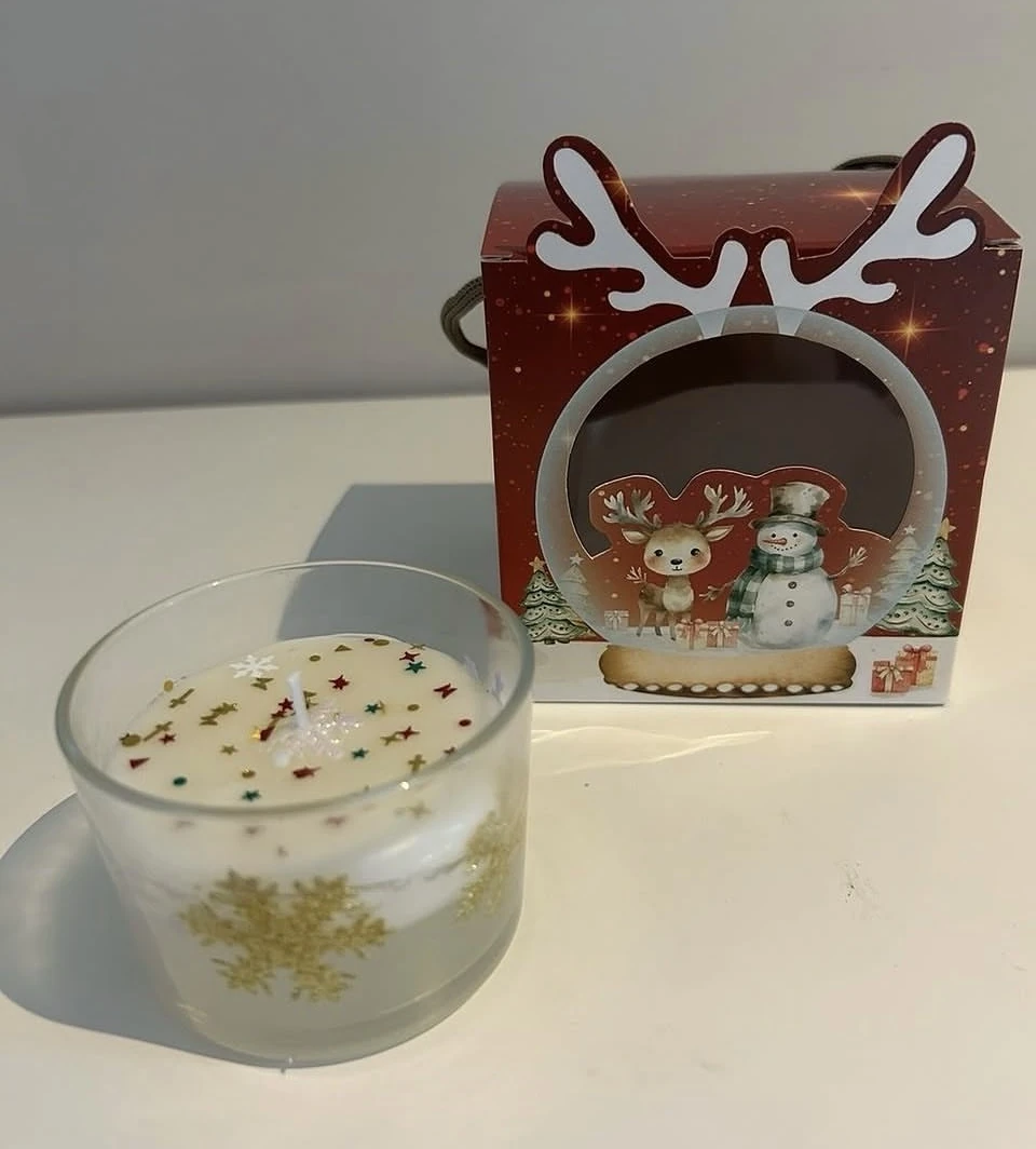 Vela em copo de vidro com flocos dourados e pequenas estrelas, ao lado de caixa com rena e boneco de neve